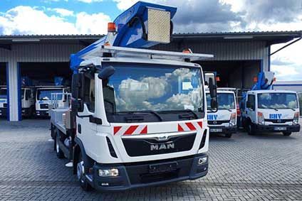Ein weißer Lieferwagen mit blauer Hubarbeitsbühne parkt vor einem Lagerhaus; weitere Servicefahrzeuge stehen in der Nähe.