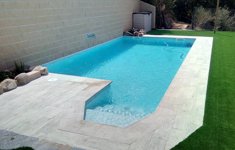 Piscina rectangular con agua azul cristalina, patio de piedra y césped verde. Una pared beige al fondo.