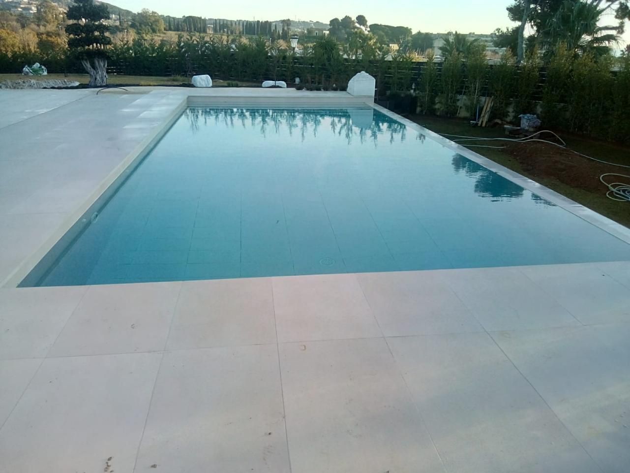 Piscina rectangular llena de agua, rodeada de hormigón de color claro, entorno al aire libre.