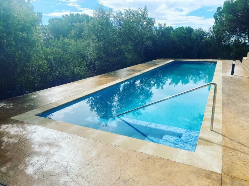 Piscina exterior rectangular de agua azul con escaleras, rodeada de hormigón claro y árboles.