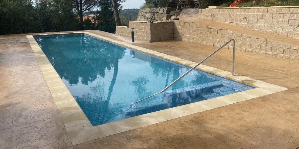 Piscina rectangular con superficie de azulejos azules y barandilla metálica. Está rodeada por un pavimento de piedra beige.