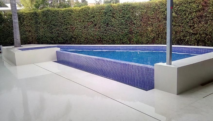 Piscina con azulejos azules, bordeada de hormigón blanco, frente a un seto verde.