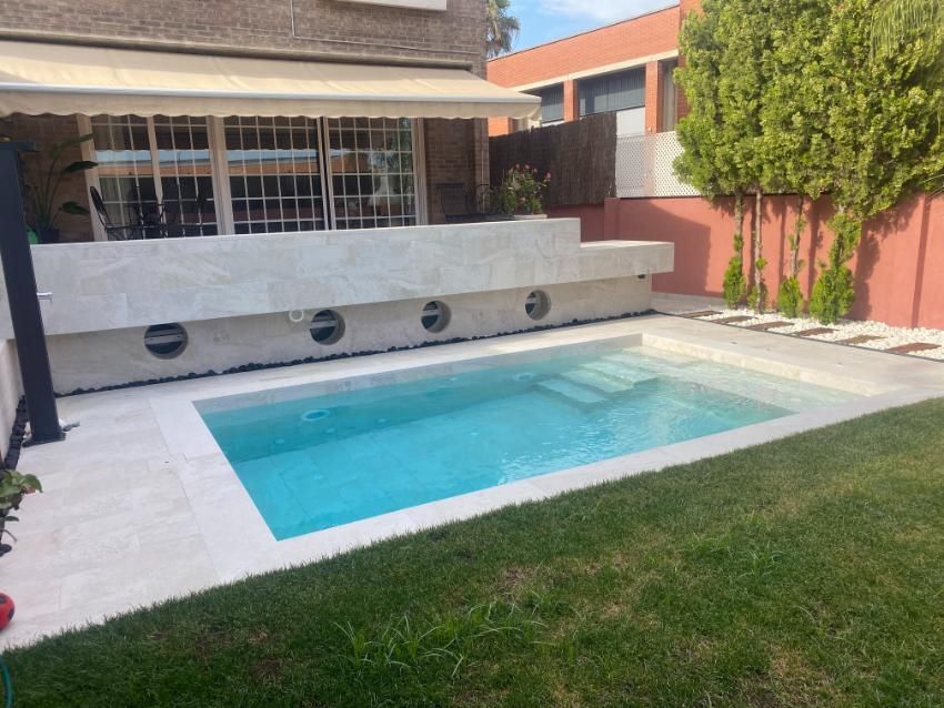 Piscina en el patio trasero con marco de piedra blanca, agua azul claro y pared blanca escalonada.