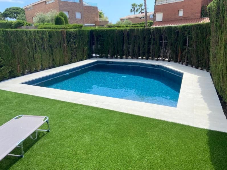 Una piscina en el patio trasero con agua azul rodeada de azulejos blancos y césped verde, con una cerca de seto.