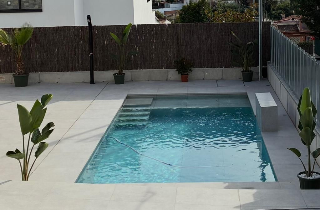 Pequeña piscina rectangular con escalones y asientos, rodeada de azulejos de color claro. Al fondo, plantas y una valla.