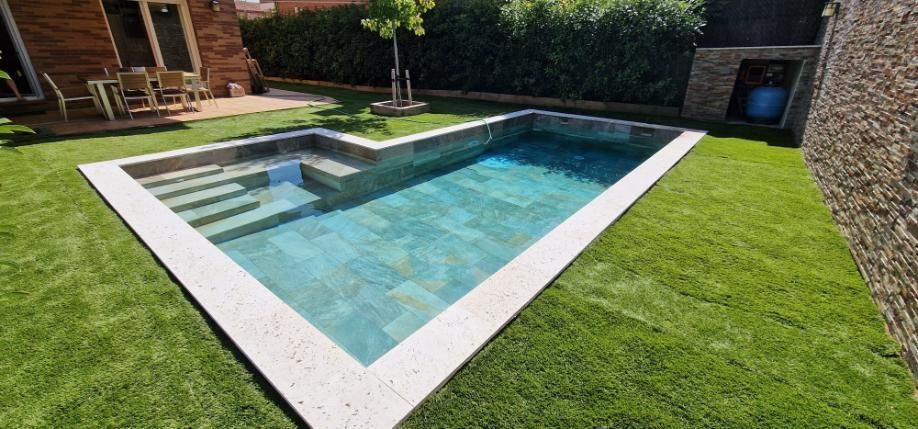 Una pequeña piscina en forma de L en un césped verde. Hay escaleras para acceder a la piscina. Se ve un edificio al fondo.