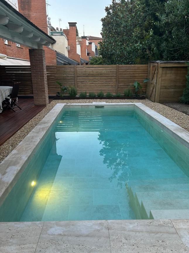 Piscina rectangular rodeada de piedra beige y pequeños guijarros, con fondo de valla de madera.