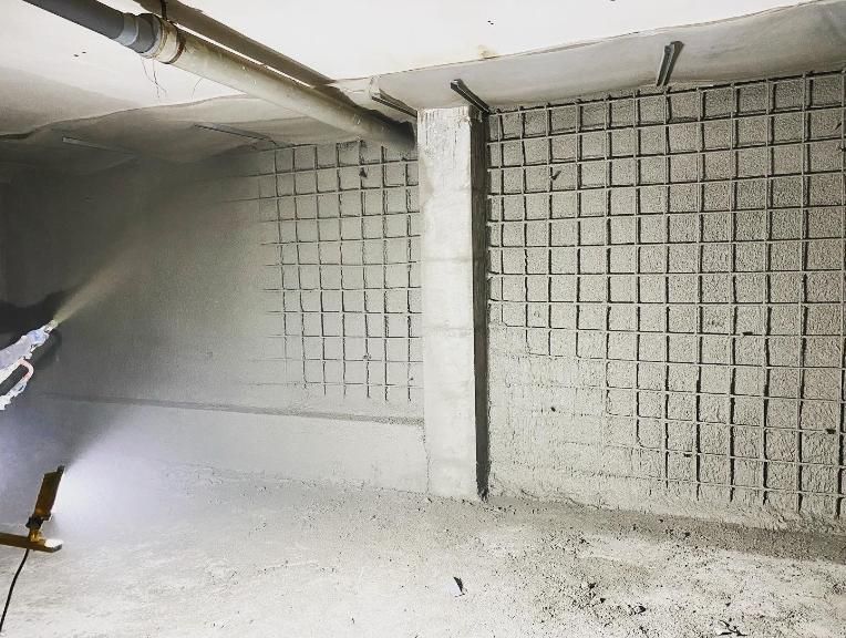 Aplicación de hormigón con pulverizador sobre una pared reforzada con rejilla en una obra de construcción interior.