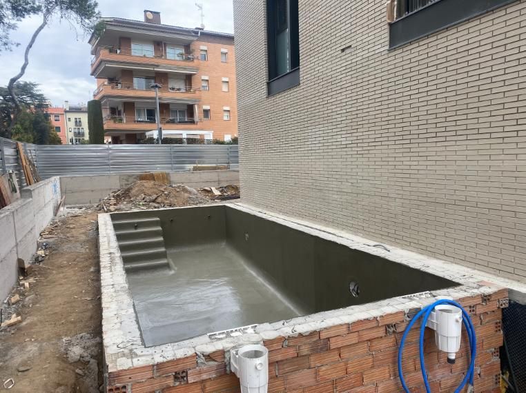 Una piscina rectangular en construcción con paredes de ladrillo y hormigón, junto a un edificio moderno.