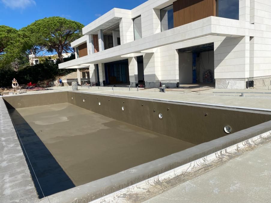 Piscina rectangular vacía en construcción frente a una casa blanca moderna con grandes ventanales.