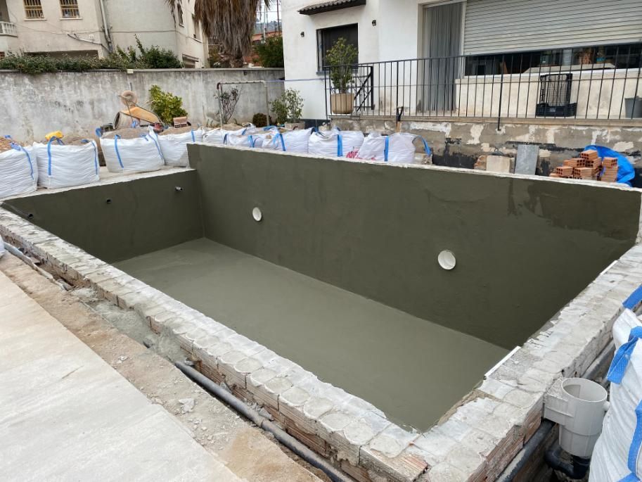 Piscina rectangular recién enlucida en construcción. Ladrillo beige y bolsas blancas la rodean.