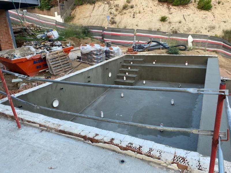 Piscina de hormigón en construcción, con escaleras y grifería. Se ven un contenedor de escombros y una ladera.