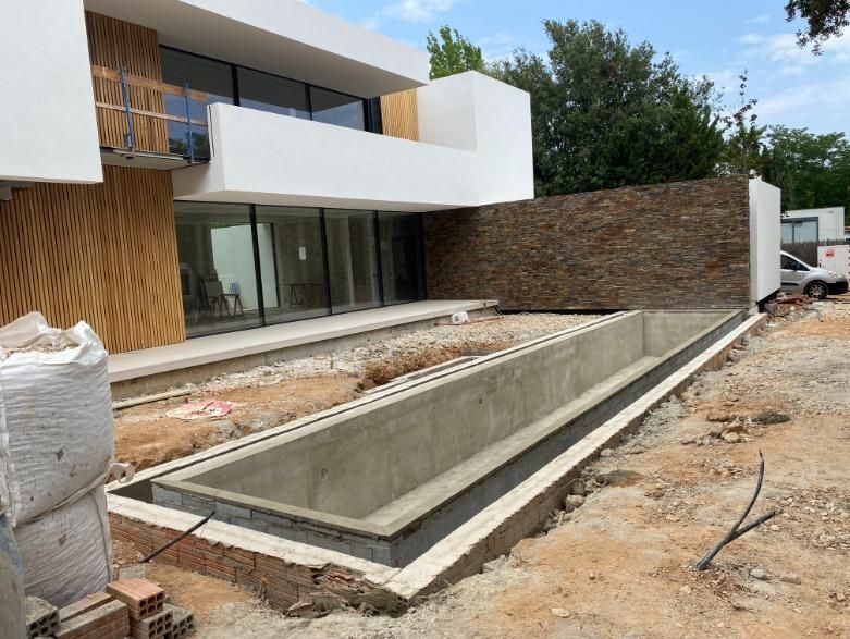 Solar en construcción con piscina rectangular parcialmente construida junto a casa moderna con revestimiento de madera