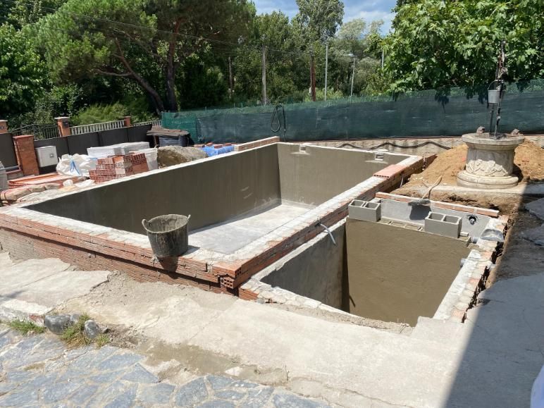 Piscina en construcción; estructura de ladrillo y hormigón al aire libre con árboles al fondo.
