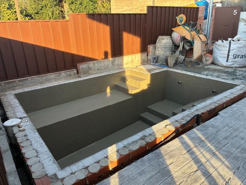 Piscina rectangular en construcción con escalones, borde de ladrillo y revestimiento de cemento. Valla de madera al fondo.
