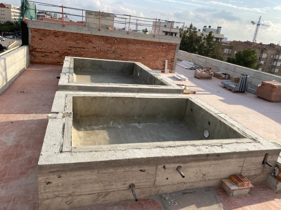 Dos piscinas de hormigón en construcción en una azotea, con una pared de ladrillo y el horizonte de la ciudad al fondo.