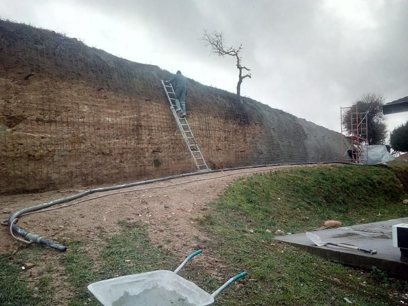 Obrero de la construcción en escalera, rociando hormigón en una ladera; manguera y carretilla en primer plano.