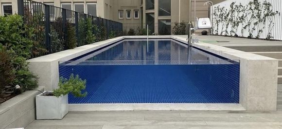 Se ve una piscina rectangular con lados de azulejos azules y paredes de cristal, con un edificio y vegetación al fondo.