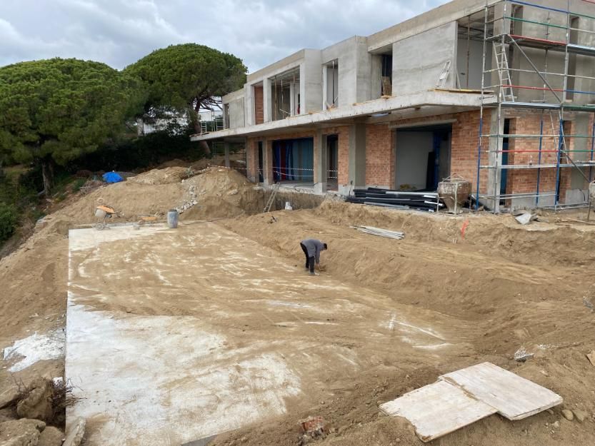 Obra con una casa a medio construir y un trabajador. Se ve maquinaria de movimiento de tierras.