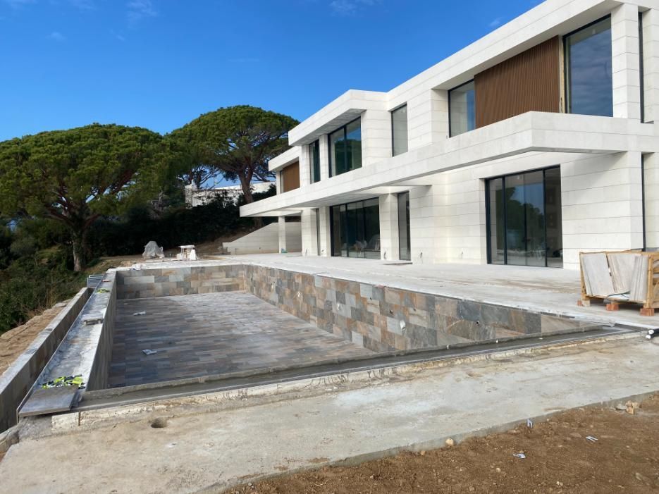 Casa moderna en construcción con piscina rectangular. Cielo azul, árboles y detalles de piedra.