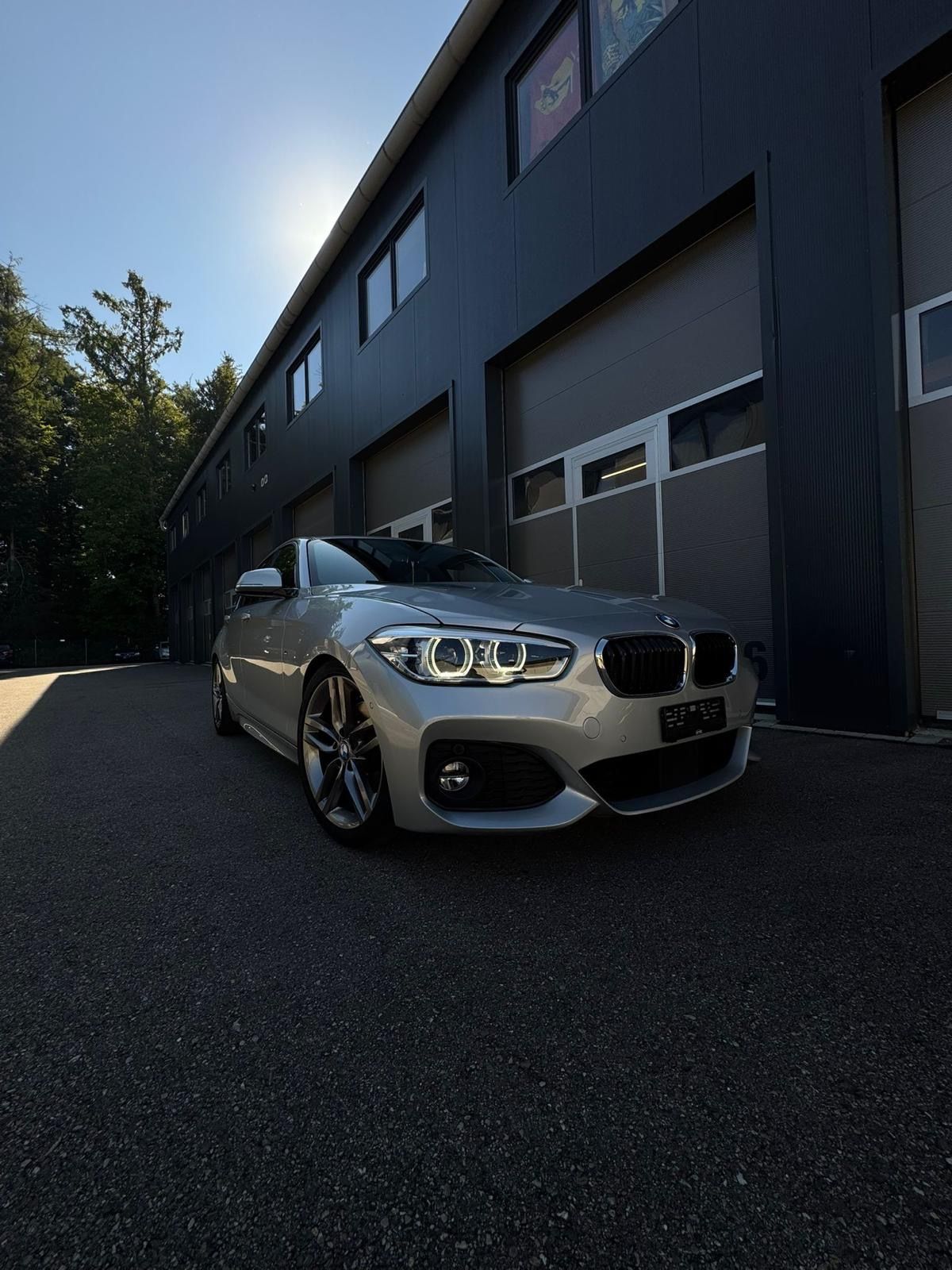 Silberner BMW 1er, geparkt vor einem modernen Gebäude mit offenen Garagentoren. Helles Sonnenlicht.