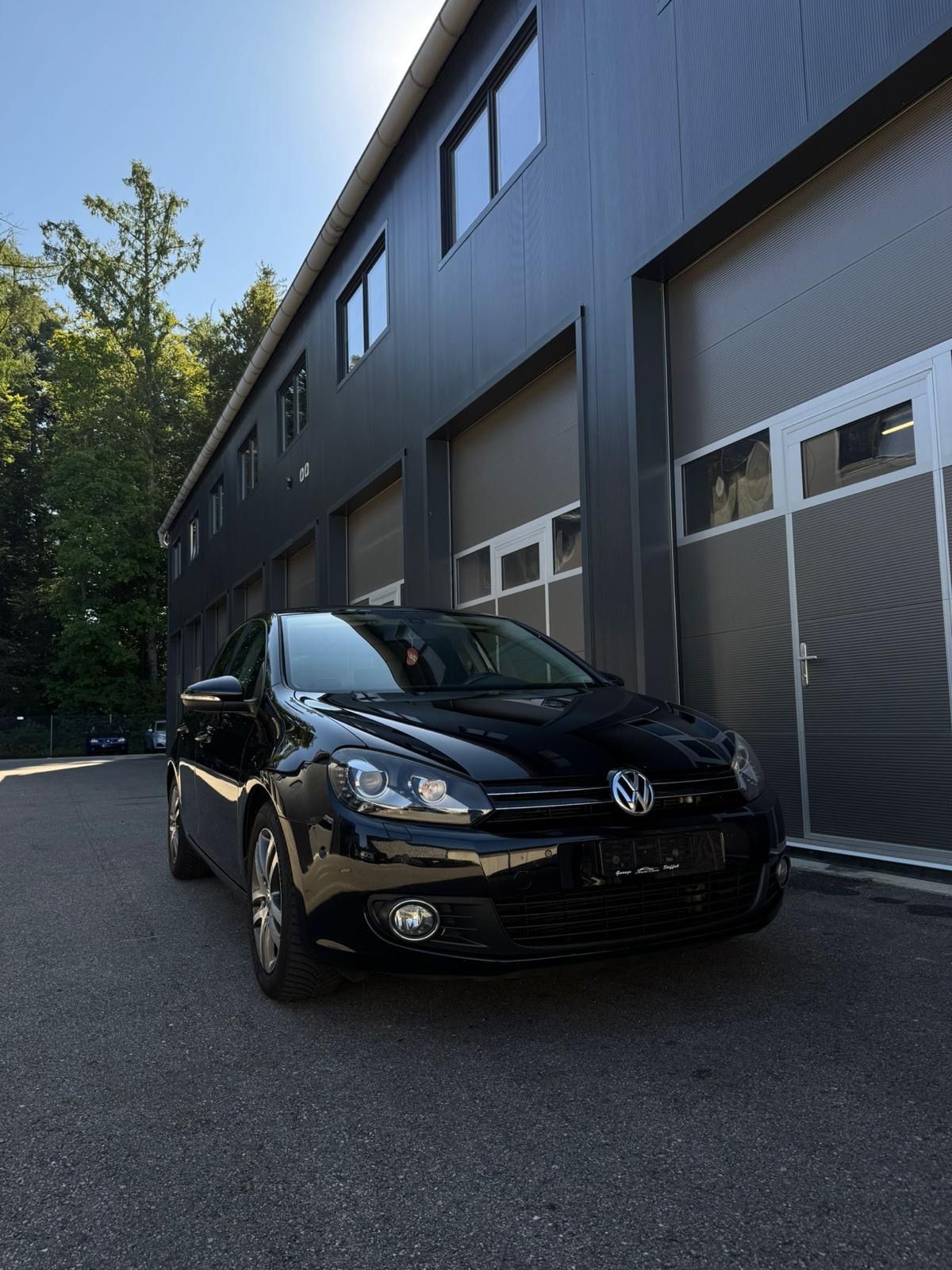 Schwarzer Volkswagen Golf, geparkt vor einem modernen Gebäude mit Garagentoren, sonniger Tag.