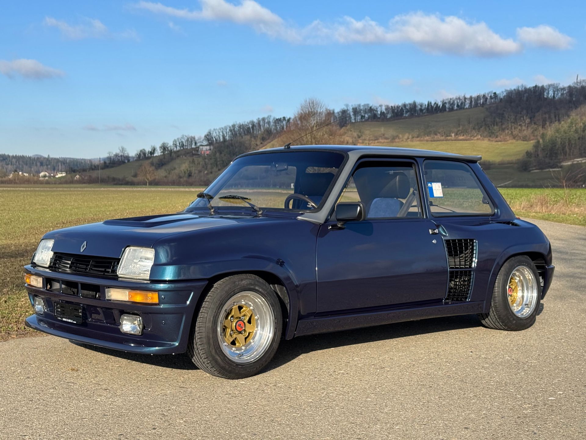 A dark blue Renault 5 Turbo sports car parked on an asphalt road in a rural landscape under a clear blue sky.