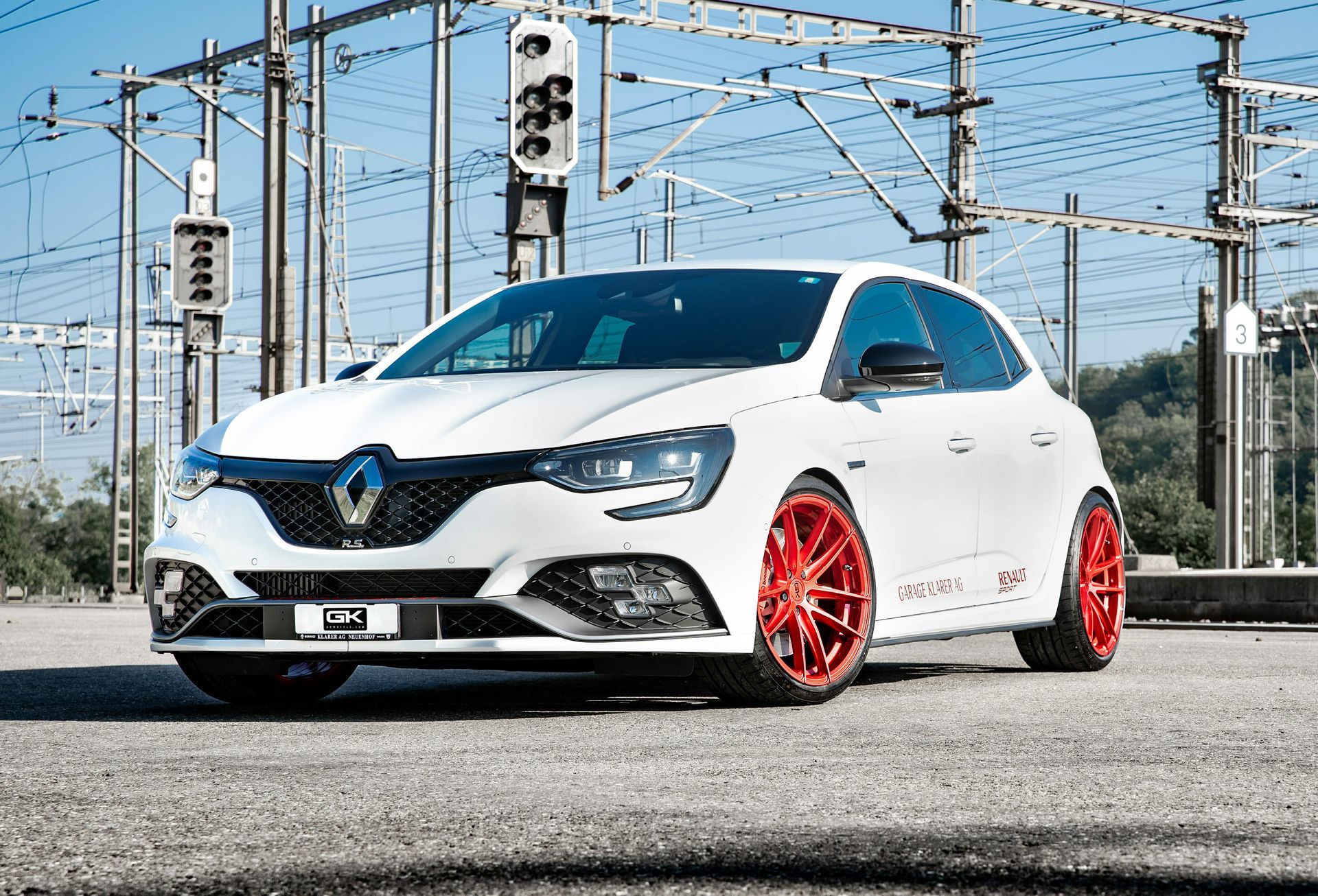 Ein weißer Renault Megane Schrägheckwagen mit roten Spezialfelgen parkt auf einem asphaltierten Parkplatz unter freiliegenden Industrie-Stromleitungen.