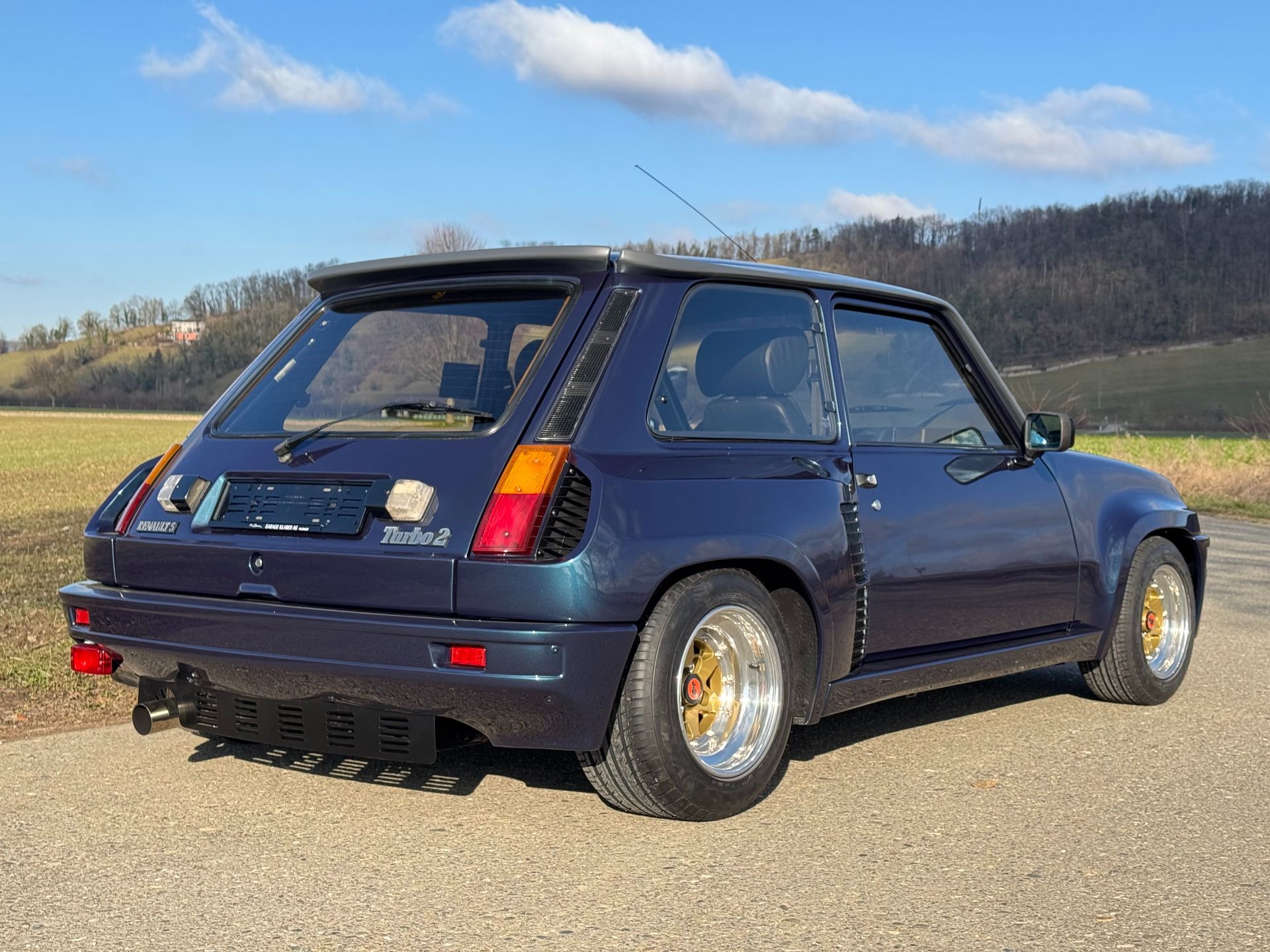 Ein dunkelblauer Renault 5 Turbo parkte auf einer asphaltierten Strasse neben einem Feld unter strahlend blauem Himmel.