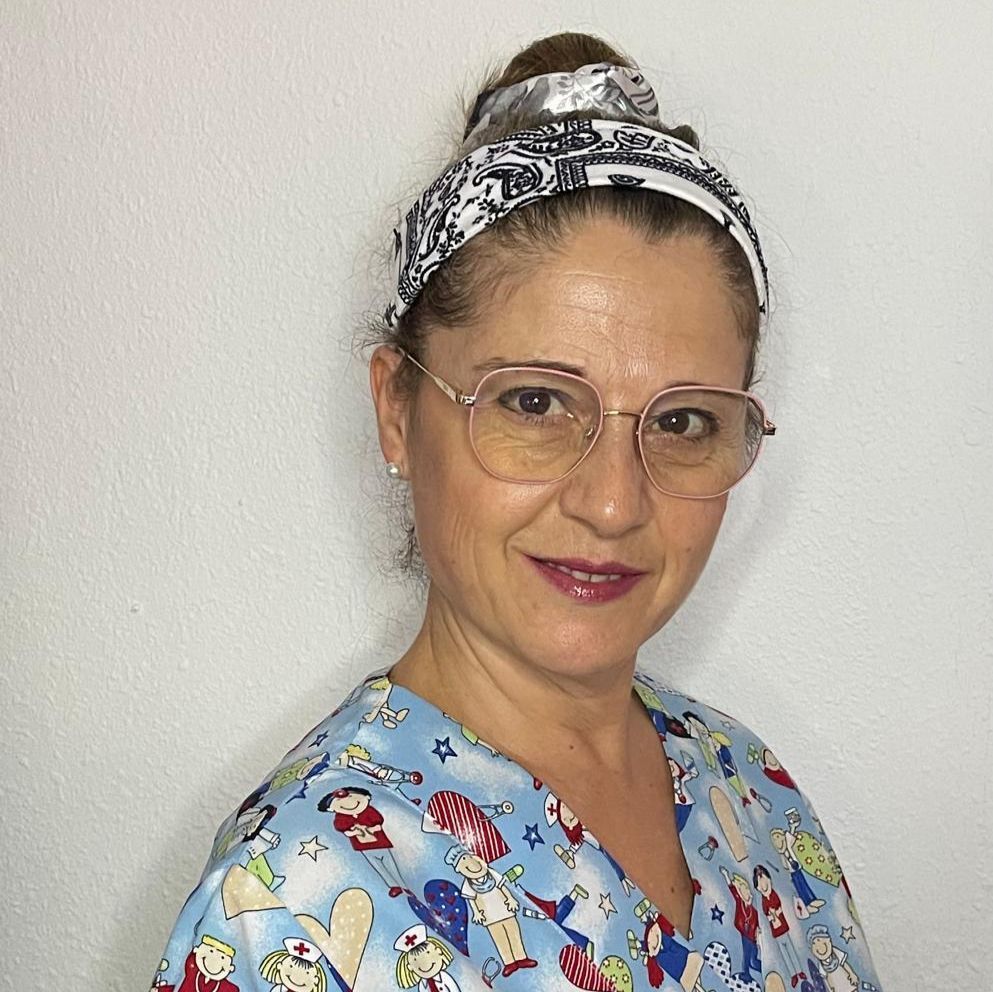 Una mujer con uniforme estampado y pañuelo en la cabeza sonríe, usando gafas, en un lugar interior.