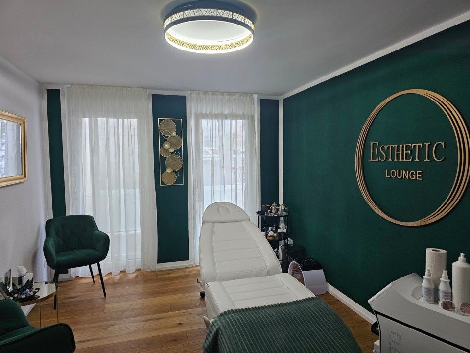 Interieur der Esthetic Lounge: grüne Akzentwand, Behandlungsliege, goldenes Logo, Fenster und zwei Stühle.