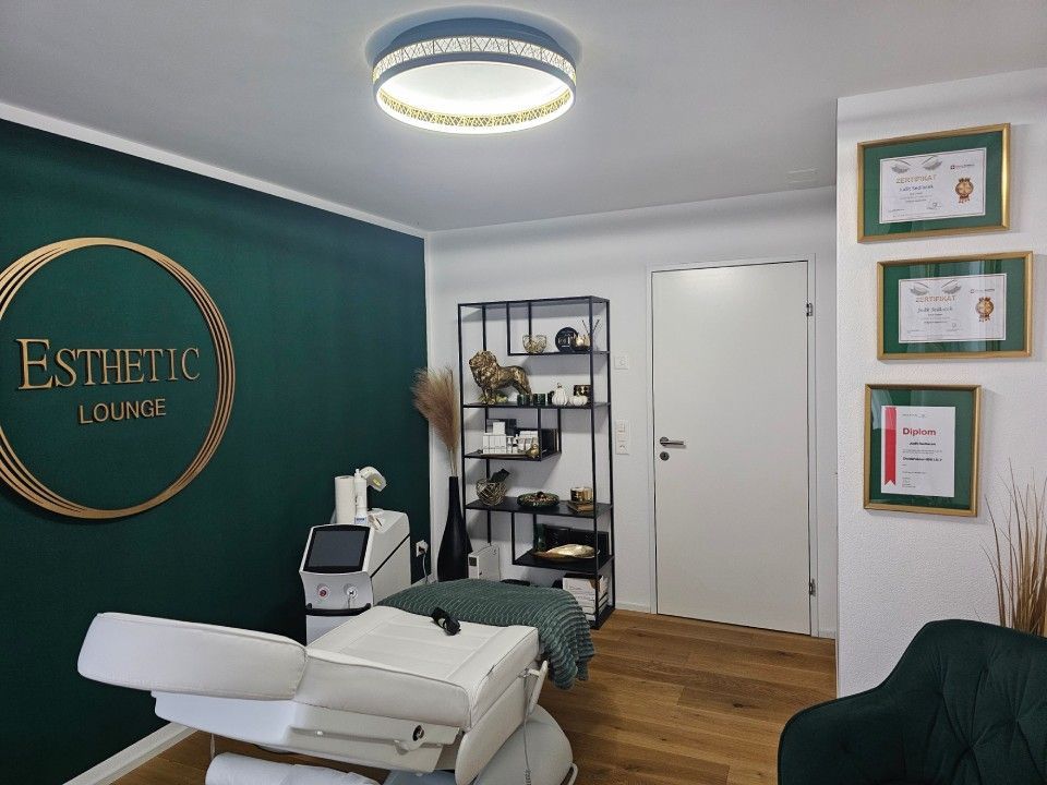 Ästhetisch gestalteter Lounge-Bereich mit Behandlungsliege, dunkelgrüner Akzentwand mit Logo,  moderner Beleuchtung.