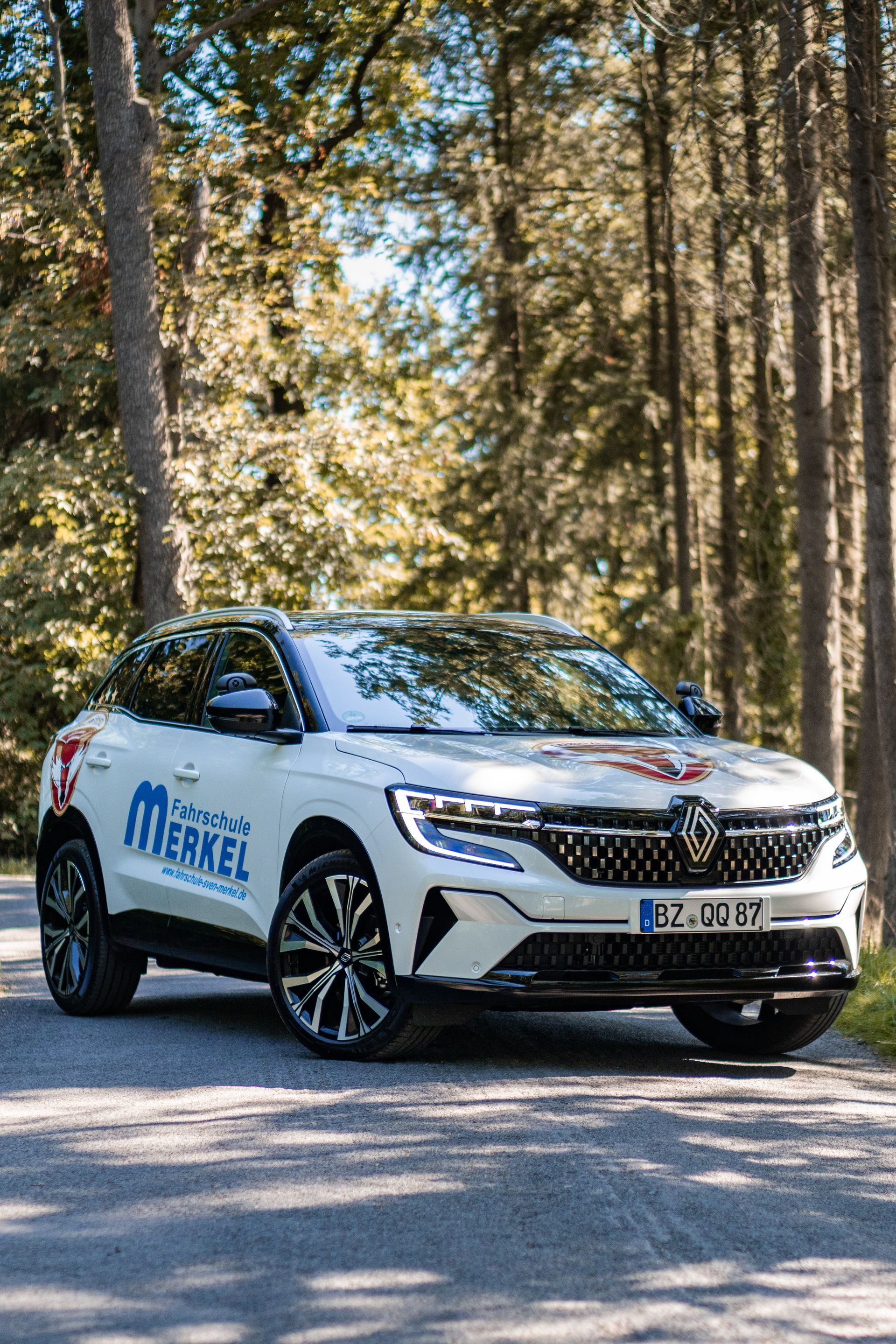 Weißer Renault-SUV mit Logo, geparkt auf einer Straße in einer Waldlandschaft, Bäume im Hintergrund.