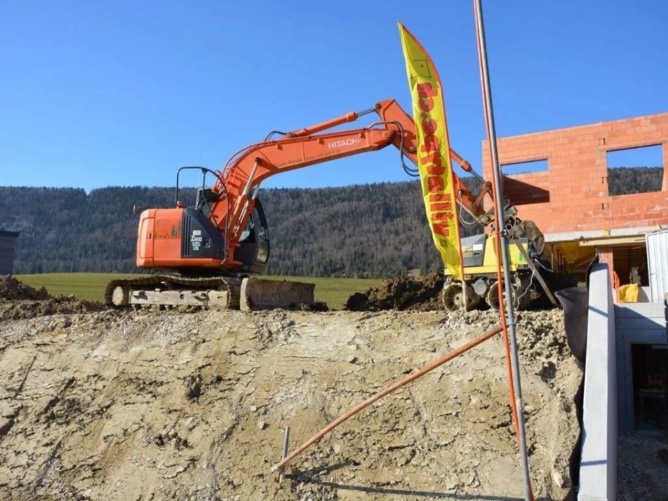 Une pelleteuse orange enlève de la terre sur un chantier de construction, avec un bâtiment en briques partiellement construit en arrière-plan.