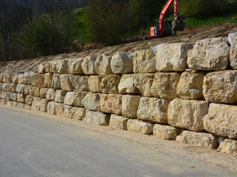 Mur de soutènement en pierre le long d'une route, construit pour stabiliser le flanc de la colline ; une excavatrice travaille au-dessus.
