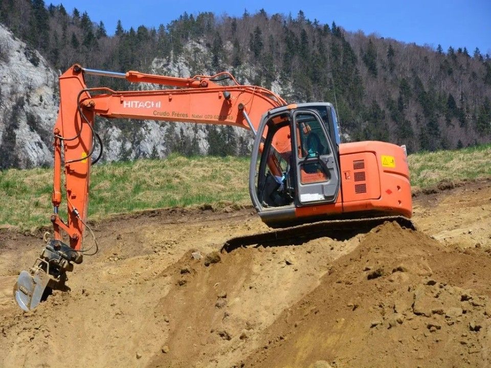 Une pelleteuse orange creuse un flanc de colline ; des arbres et le ciel se dessinent à l'arrière-plan.