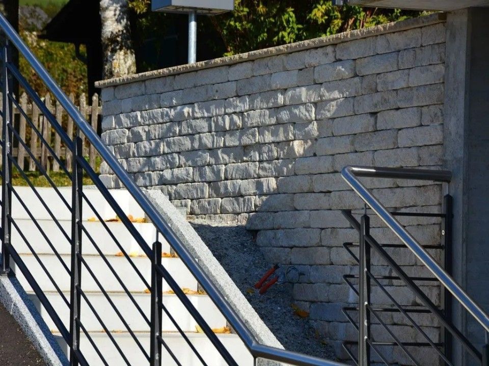 Escalier à rampe métallique montant vers un mur de pierre ; la lumière du soleil projette des ombres.