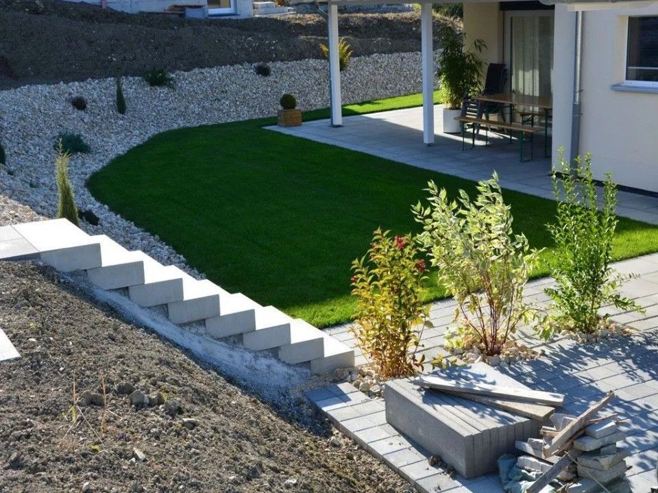 Un jardin en terrasses avec des marches, une pelouse verte, un mur de soutènement en gravier, une terrasse et des arbustes.