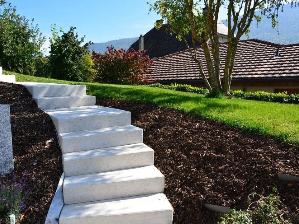 Des marches en béton montent un flanc de colline recouvert de paillis dans un jardin ensoleillé, menant vers une maison.