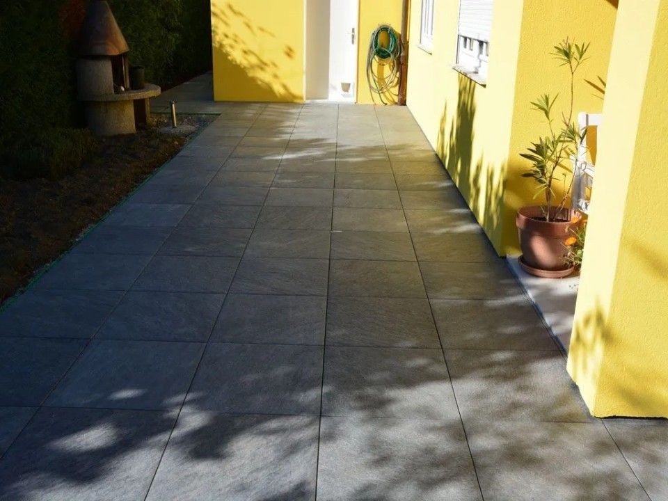 Terrasse carrelée grise devant un bâtiment jaune, avec des ombres et une plante en pot.