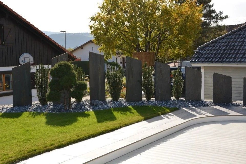 Jardin avec dalles de pierre dressées, arbustes et galets gris sur une pelouse verte près d'une maison.