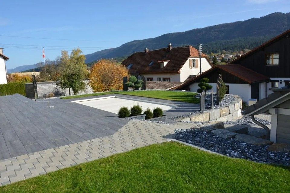 Quartier résidentiel avec terrasse pavée, pelouse verdoyante et maisons se détachant sur un fond montagneux par une journée ensoleillée.