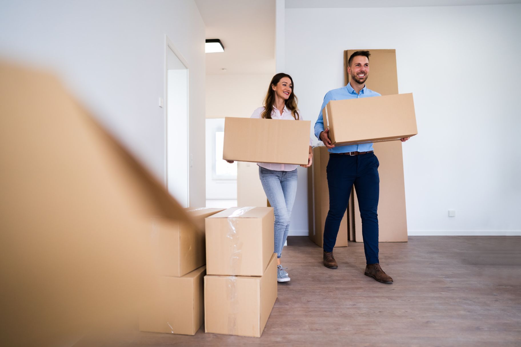 Pareja sonriente cargando cajas de cartón a su nueva casa. Se ven cajas y paredes desnudas.