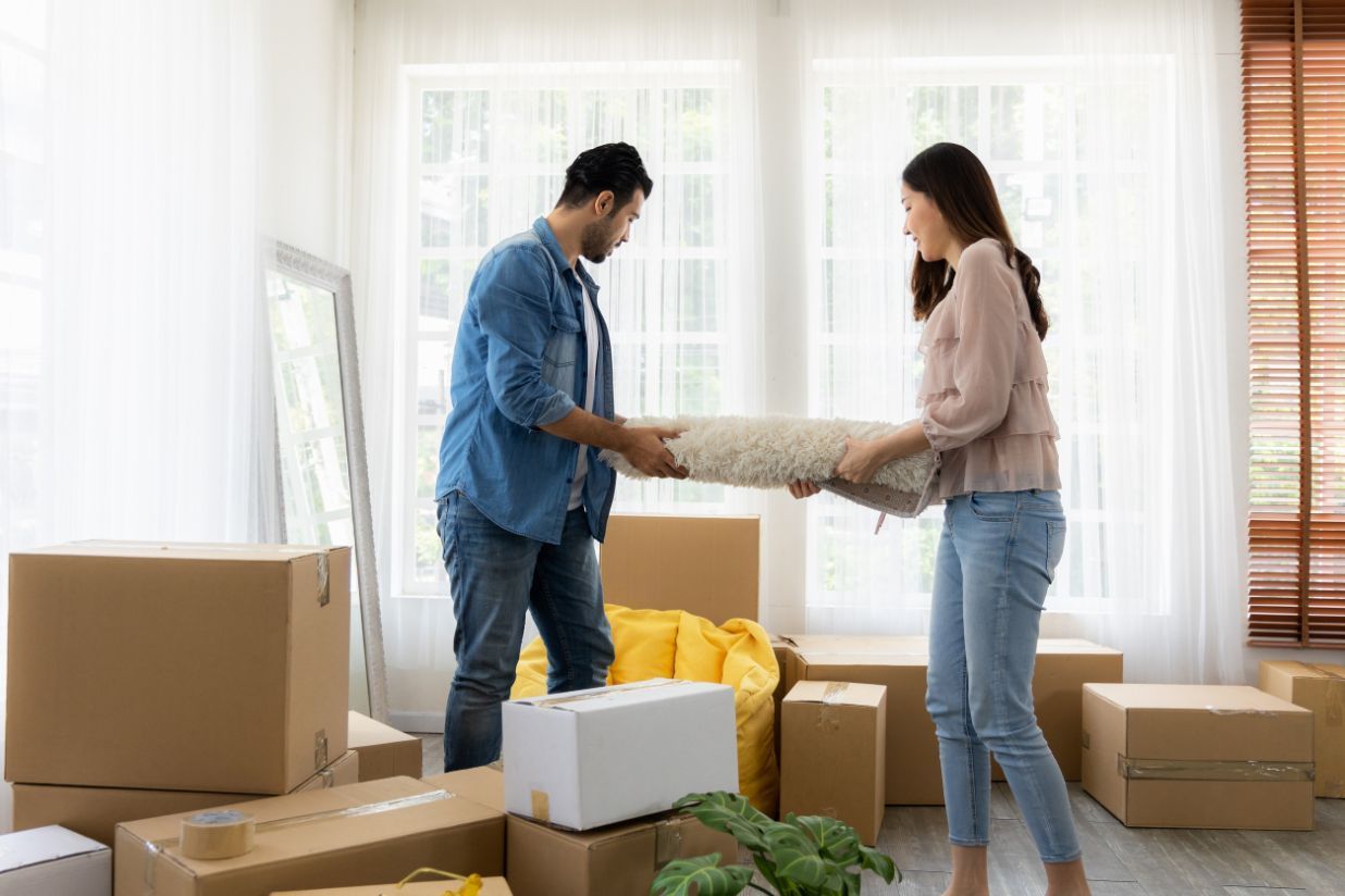 Pareja desenrollando una alfombra, rodeada de cajas de mudanza en una nueva casa.