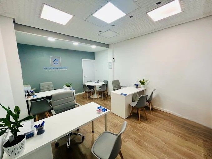 Interior de oficina con escritorios blancos, sillas grises y suelo de madera clara. Una pared luce un logotipo.