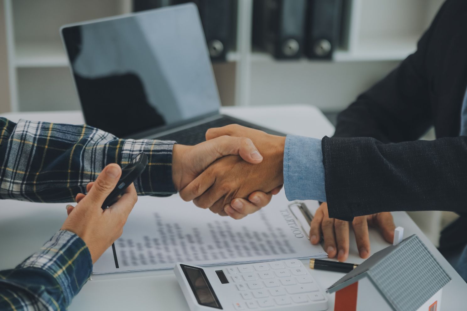 Dos personas se dan la mano sobre un contrato y una maqueta de casa en un escritorio; una computadora portátil en el fondo.
