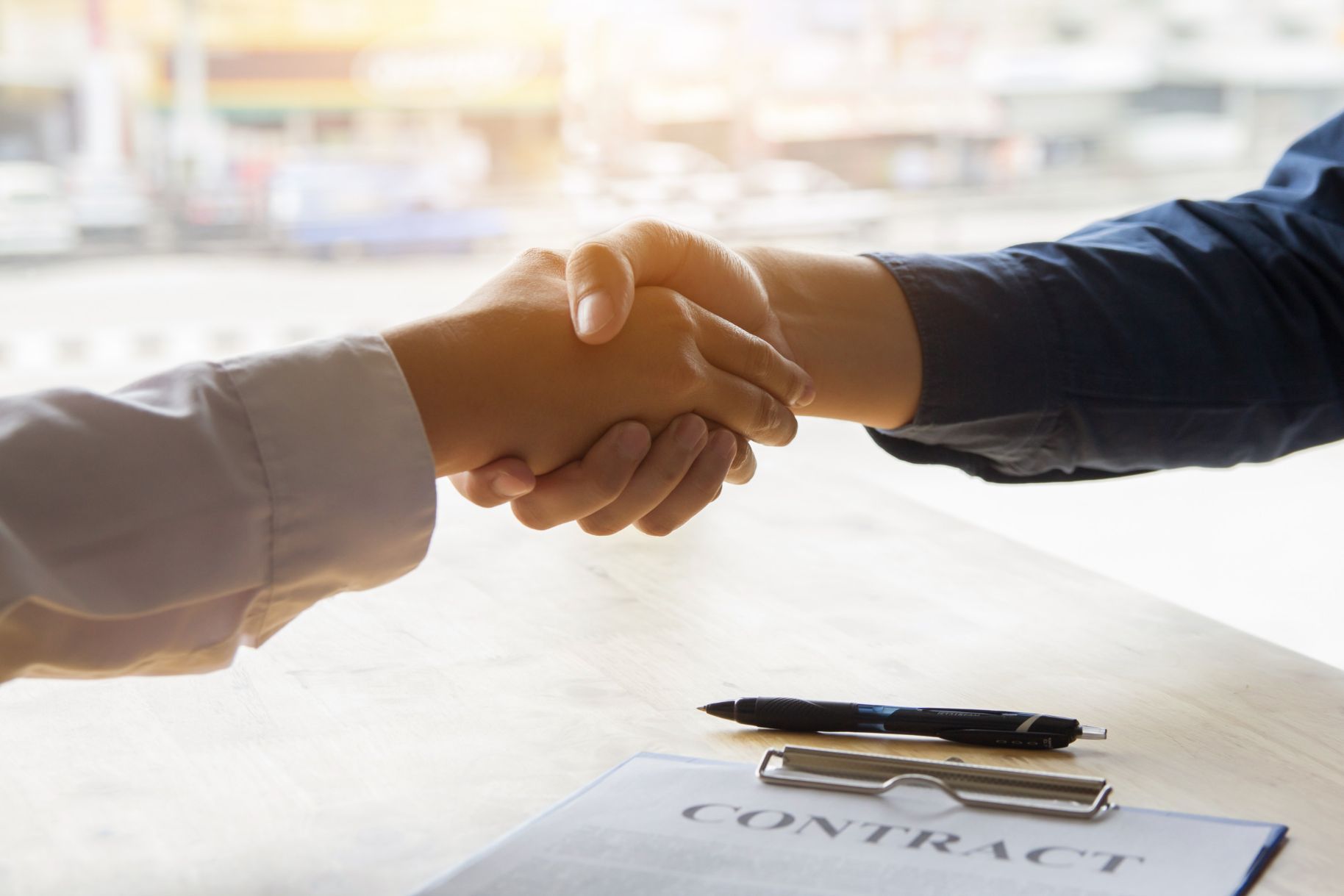 Dos personas se dan la mano sobre un contrato sobre una mesa; un bolígrafo cerca.