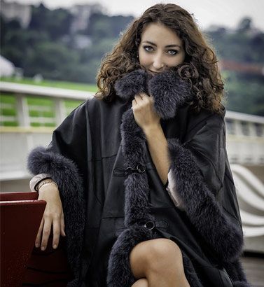 Femme vêtue d'un manteau noir bordé de fourrure, assise à l'extérieur, souriant à l'objectif.