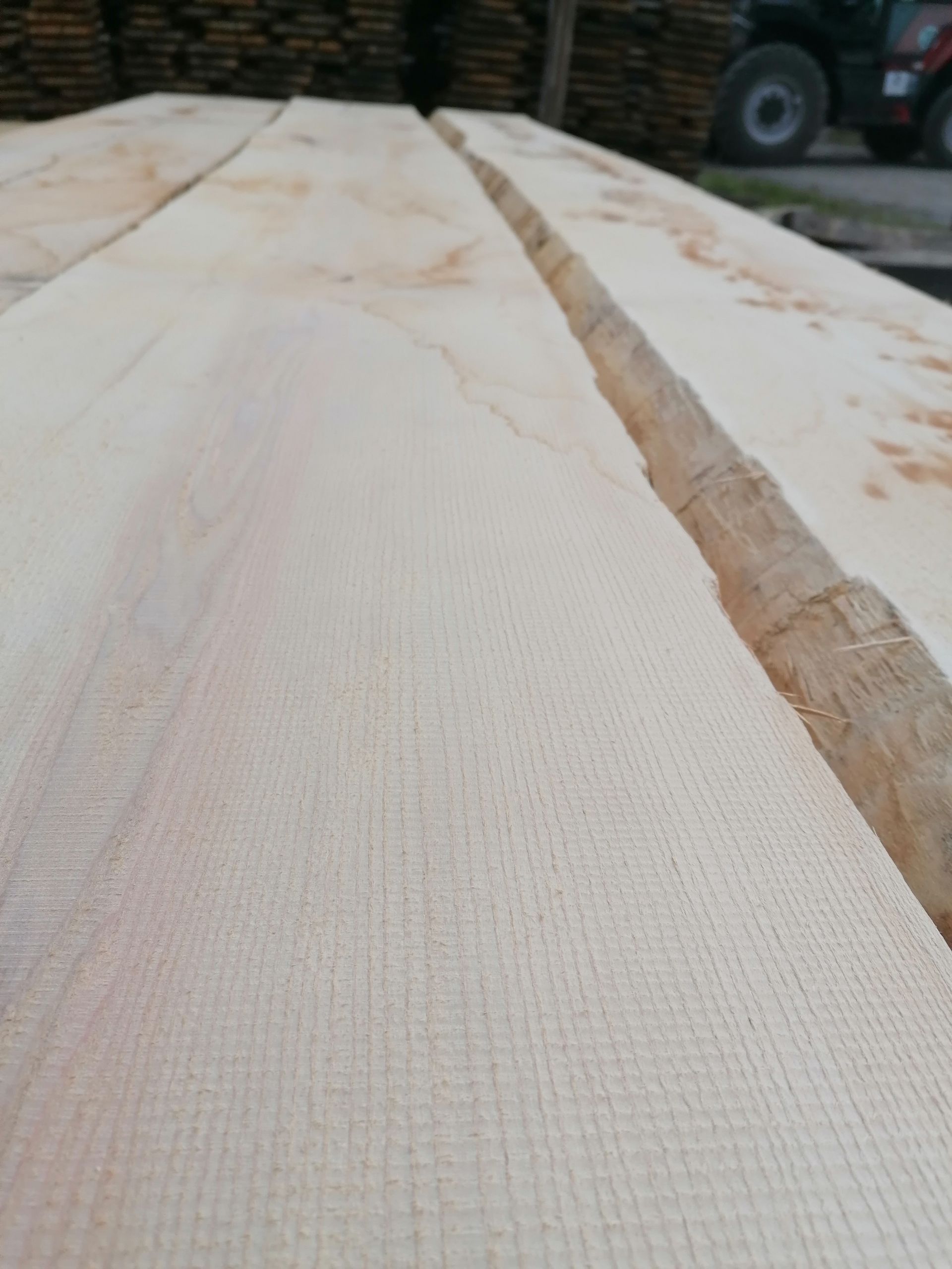 Nahaufnahme von draußen gestapelten hellen Holzbrettern, im Hintergrund ein Traktorreifen.