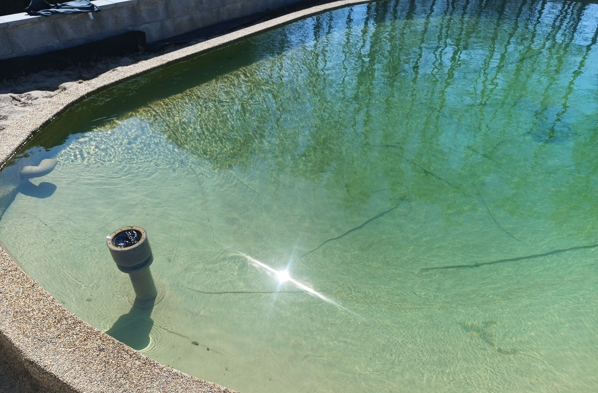 Ein flacher, türkisfarbener Teich mit einem Springbrunnen, auf dem sich das Sonnenlicht spiegelt, und einer Steinumrandung.